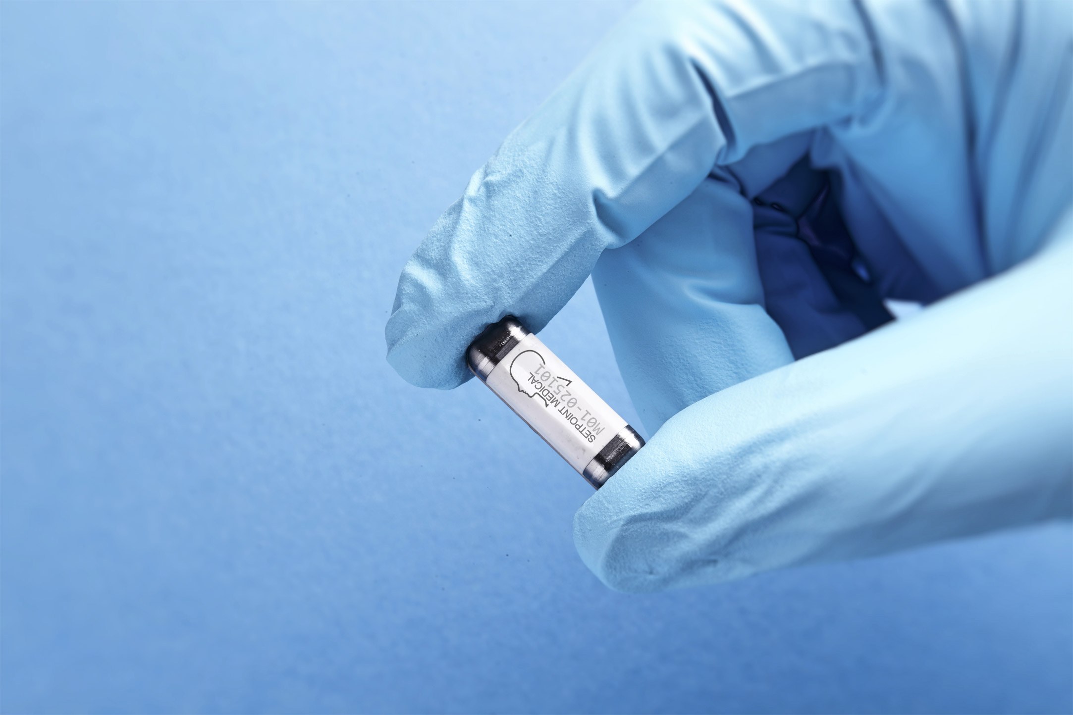 A gloved hand holding the SetPoint System implant, a small metal cylinder about the size of a vitamin capsule